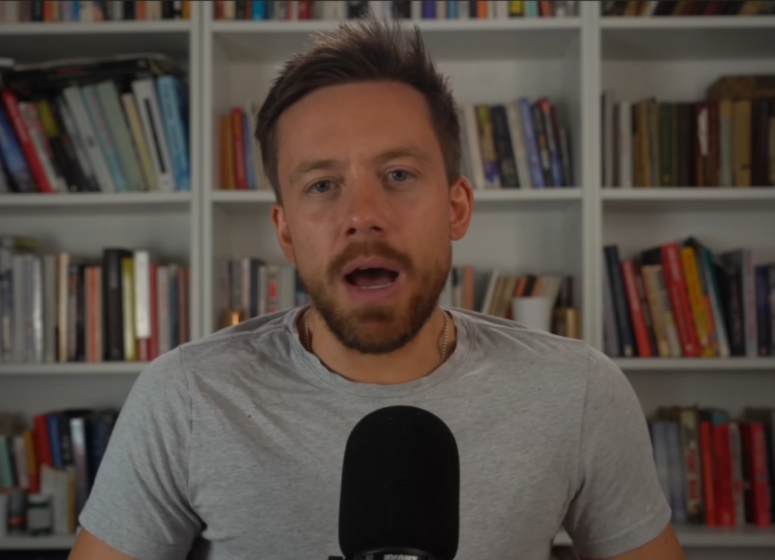 Political commentator Owen Jones speaks into a microphone during a YouTube video recorded in front of bookshelves, discussing concerns about the future of British democracy.