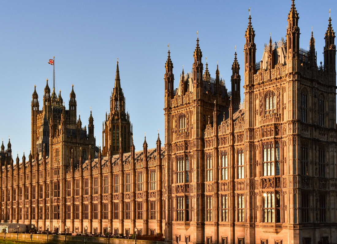 The Palace of Westminster, home to the UK Parliament, stands beside the River Thames in central London. The Gothic Revival building is illuminated by warm sunlight, with its ornate towers, detailed stonework and the Union flag visible against a clear blue sky.