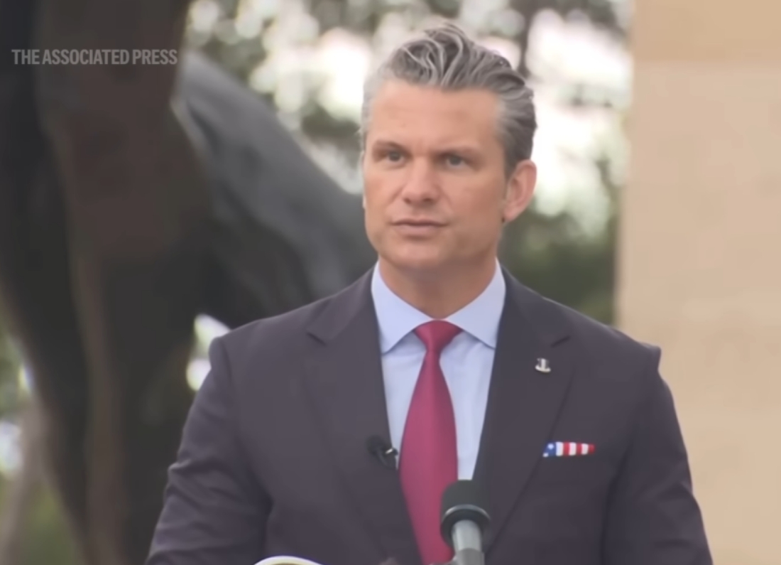 The image shows Pete Hegseth standing outdoors behind a microphone while delivering a formal speech. He is dressed in a dark suit with a light blue shirt and red tie, with an American-themed pocket square visible in his jacket. The background is softly blurred, focusing attention on Hegseth as he addresses an audience. The Associated Press watermark appears in the top-left corner. The image accompanies reporting on comments made during a D-Day anniversary commemoration event.