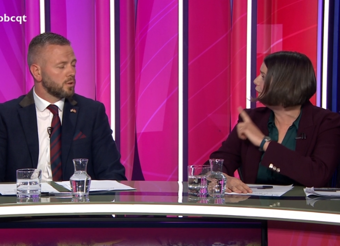 A man and a woman take part in a heated discussion during a BBC Question Time panel debate, seated behind a desk with glasses of water in front of them.