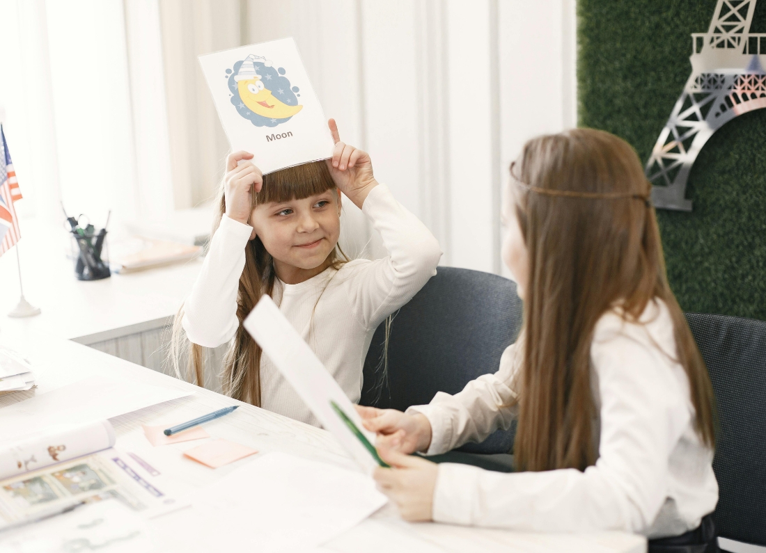 Two young schoolgirls sit at a classroom table taking part in a language-learning activity. One child holds a flashcard labelled “Moon” above her head while the other looks on, with British and American flags visible in the background.