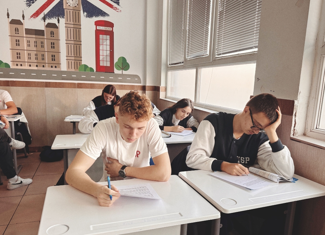 Students sitting at desks and writing in a classroom during an exam or study session, with a London-themed wall mural in the background.