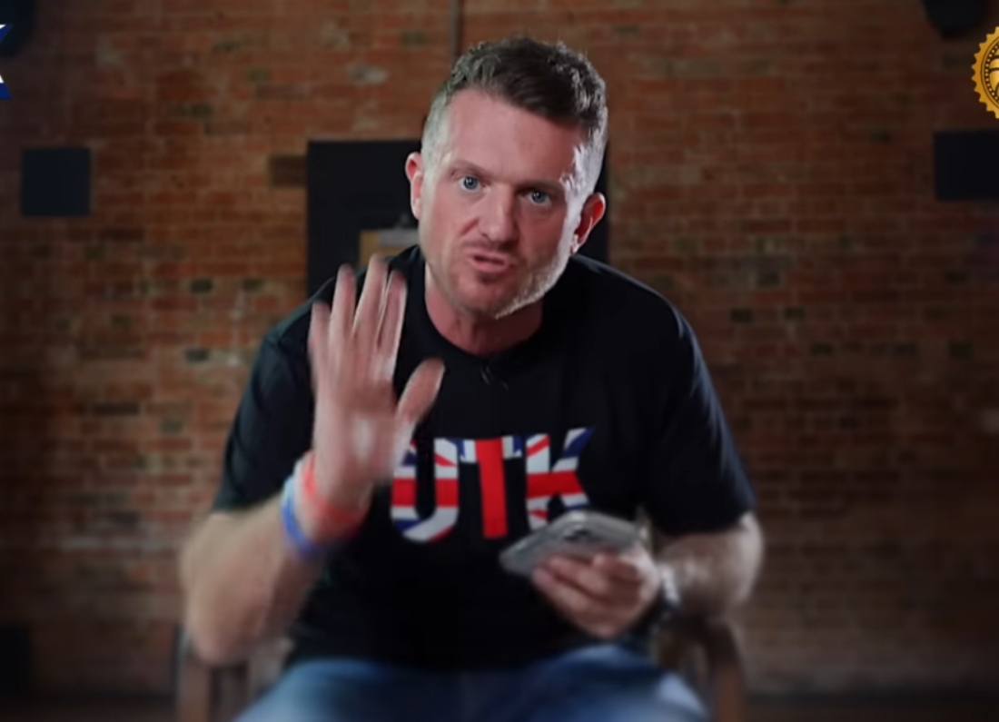 Far-right activist Tommy Robinson sits indoors holding a smartphone and gesturing towards the camera while speaking in a video recording.