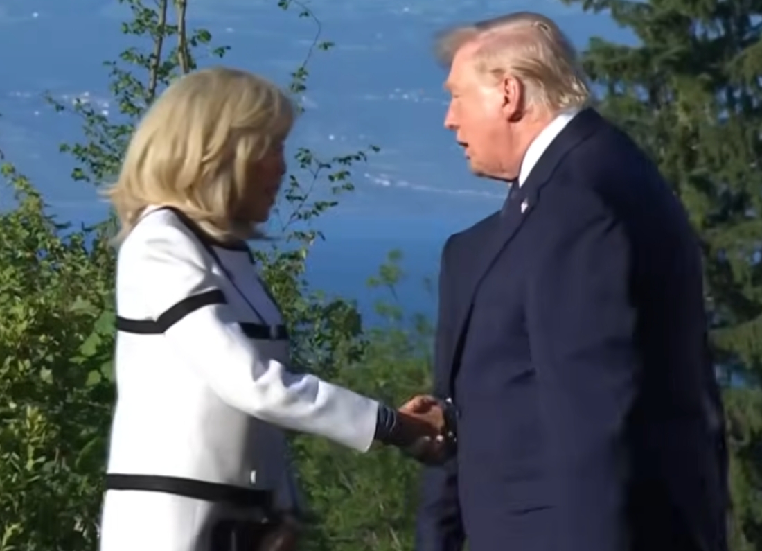 Donald Trump and Brigitte Macron shake hands outdoors during a diplomatic event, with trees and water visible in the background.