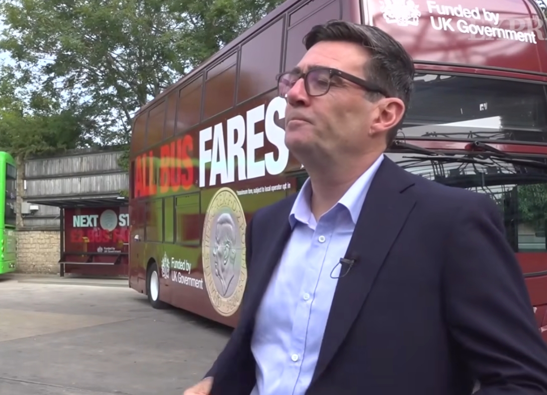 Prime Minister Andy Burnham speaks to reporters beside a government-funded bus promoting reduced fares.