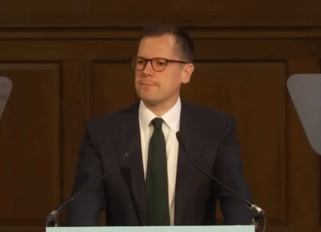 Robert Jenrick standing behind a lectern during a press conference, wearing a dark suit, white shirt and green tie.